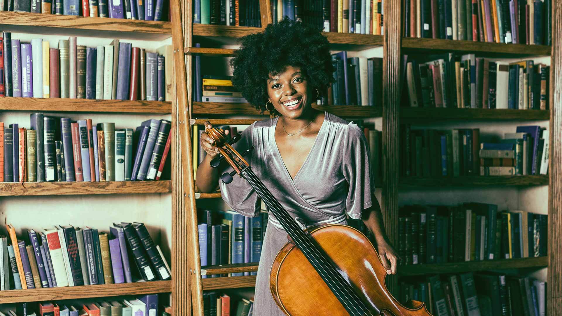 Titilayo Ayangade with her cello in front of bookshelves filled with books.