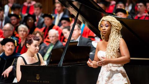  a UMD student wearing a formal gown and singing in front of a Steinway grand piano played by another UMD student. Behind them sit faculty and students donning graduation gowns. 
