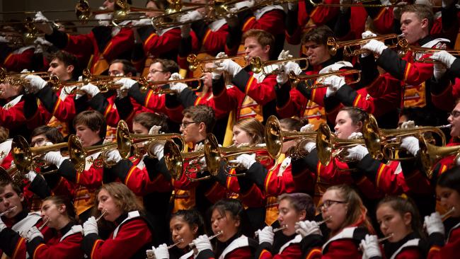  marching band members in rows wearing red, black, gold, and white uniforms and white gloves as they play various horns.